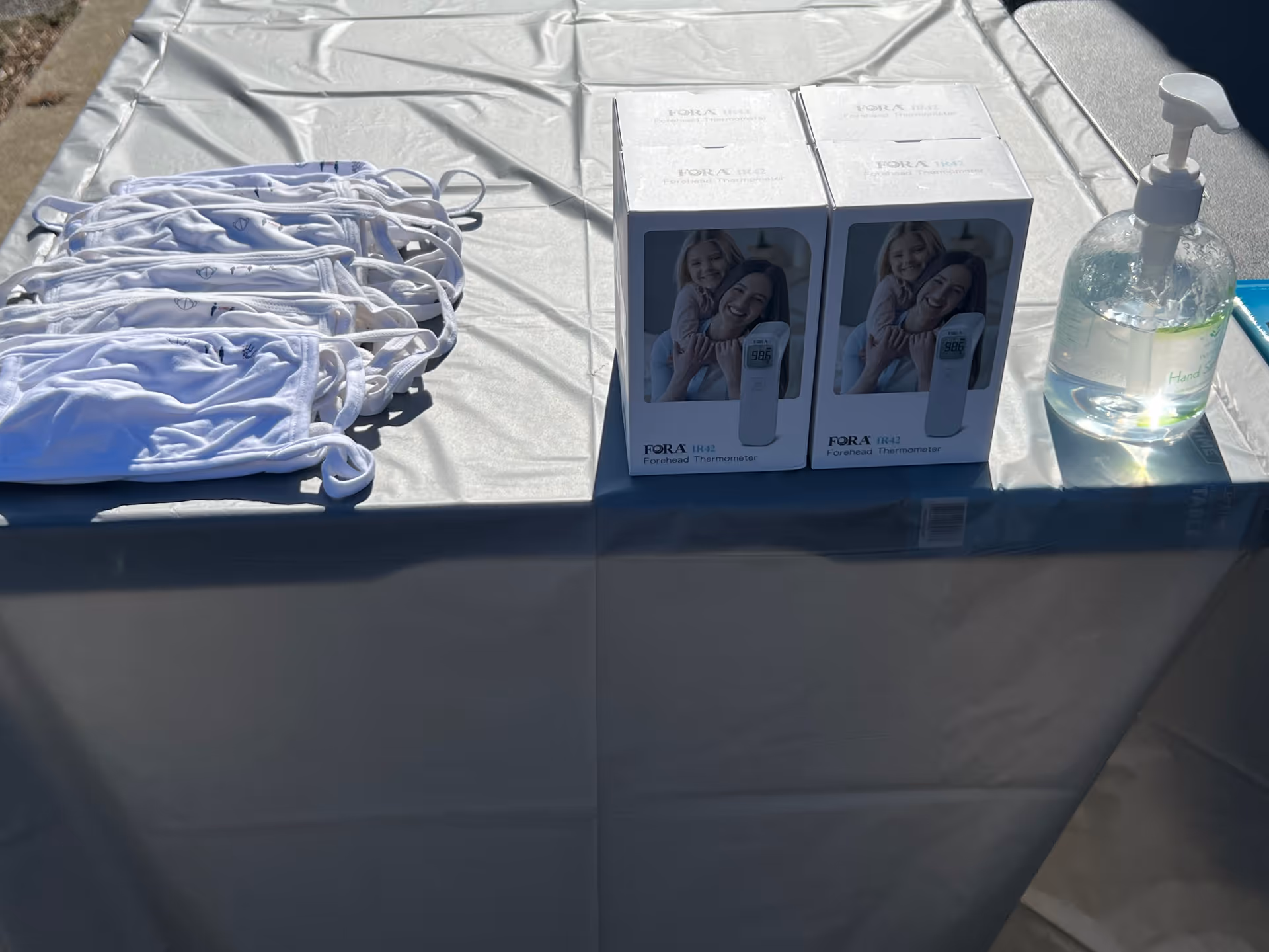 Outdoor table with white masks, thermometer boxes, and a hand sanitizer bottle.