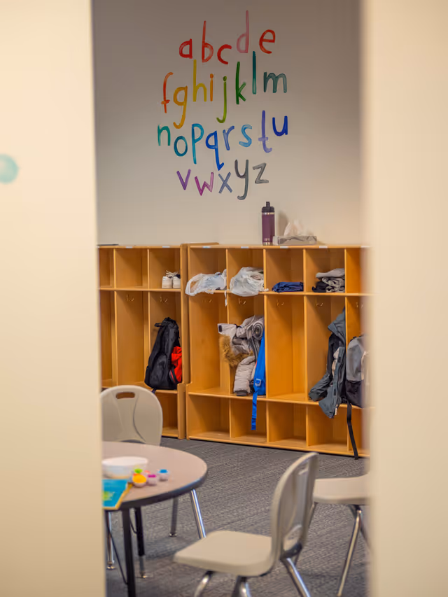 Open activity room with cubbies, jackets, a table, and colorful alphabet wall.