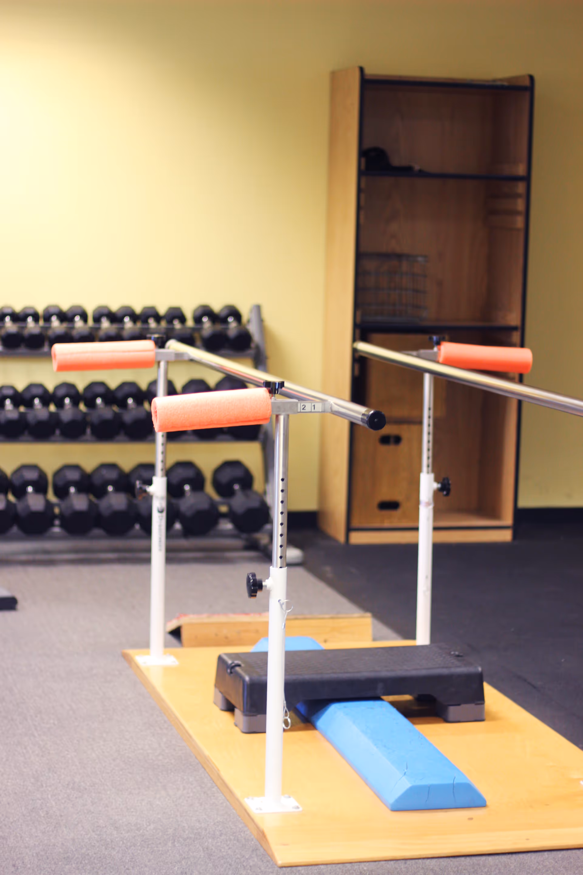 Indoor rehab gym with parallel bars, mats, and weights on a rack.