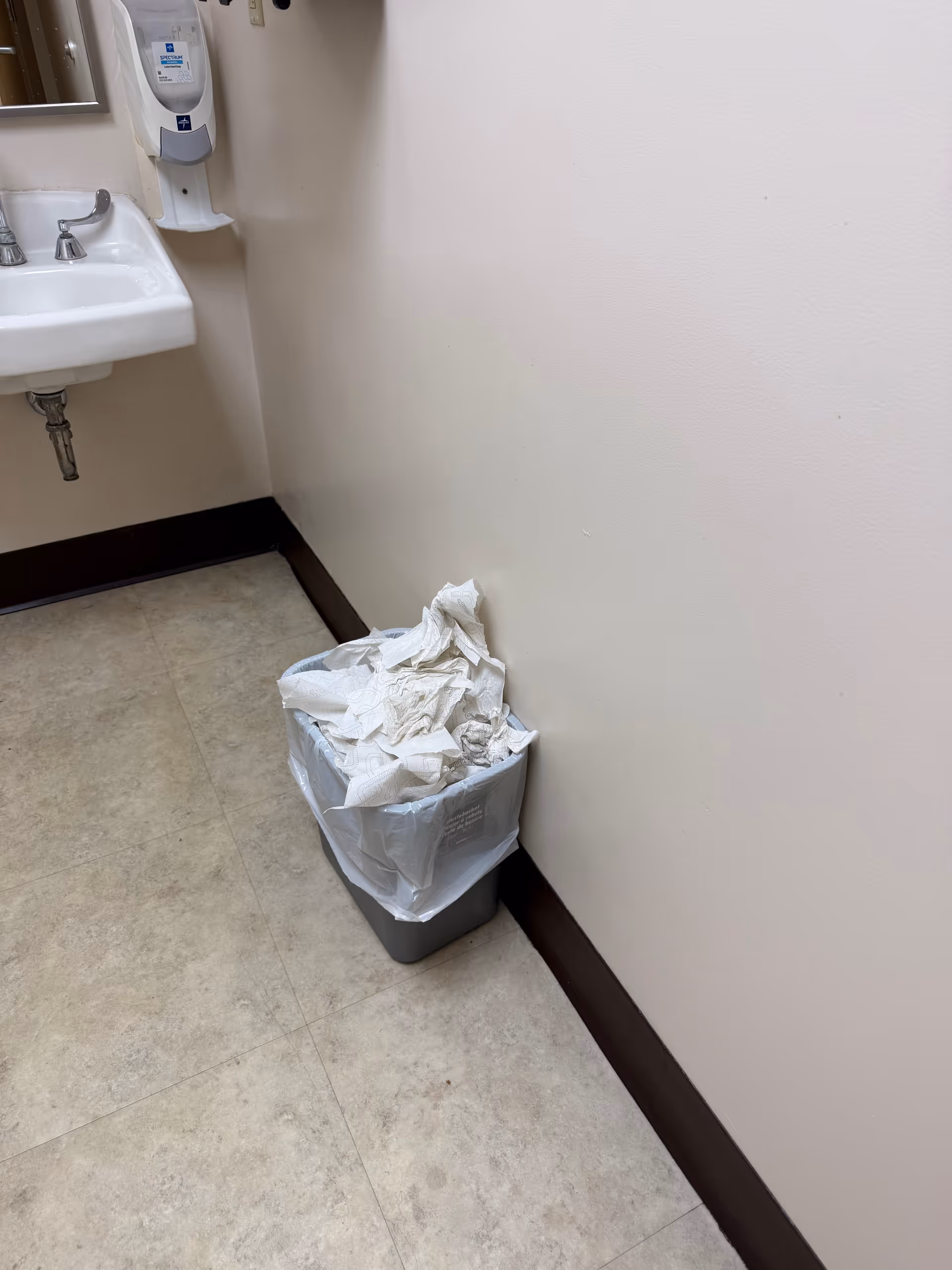 Bathroom corner with sink, soap dispenser, and trash can overflowing with crumpled paper.