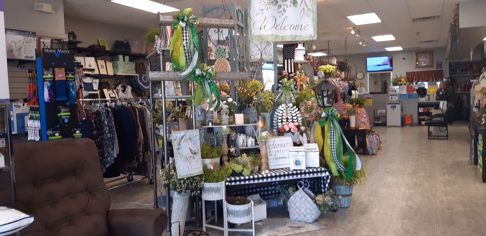 Indoor gift shop area with clothing racks, plants, decor, and a welcoming sign inside a facility.