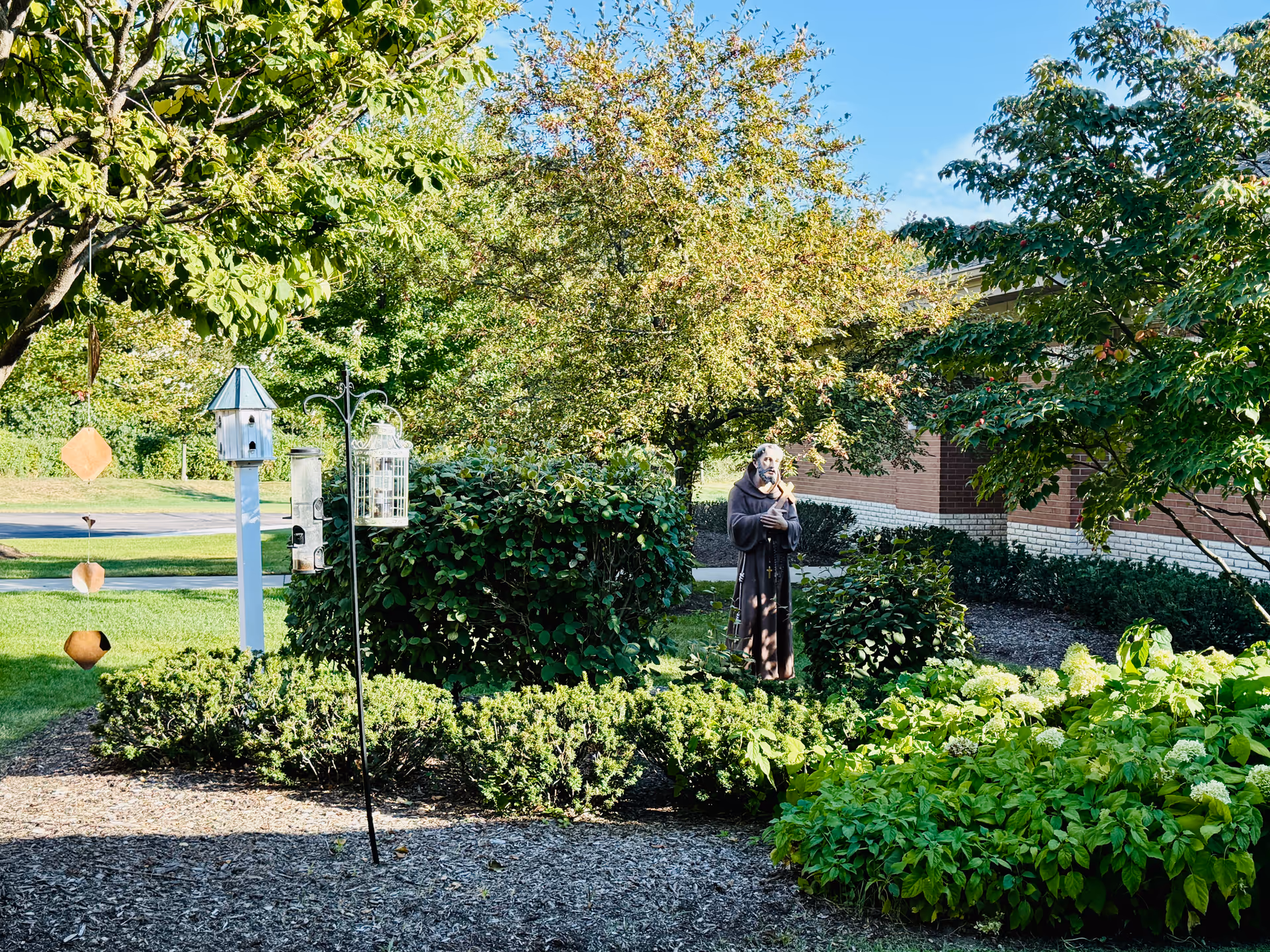 Outdoor garden area with trees, hedges, birdhouses, and a statue near a brick building.