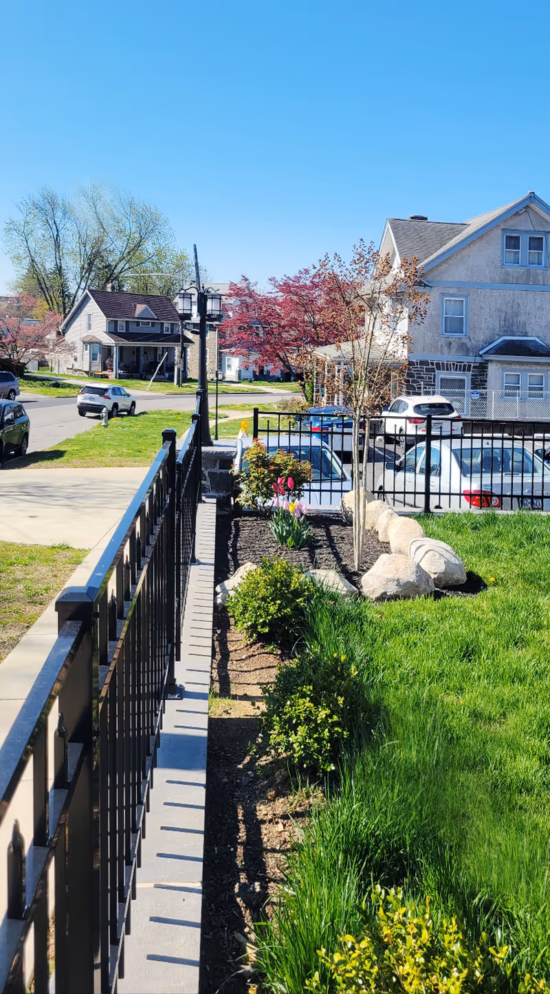 Sunny outdoor scene of a landscaped walkway with railings, trees, and nearby parked cars.