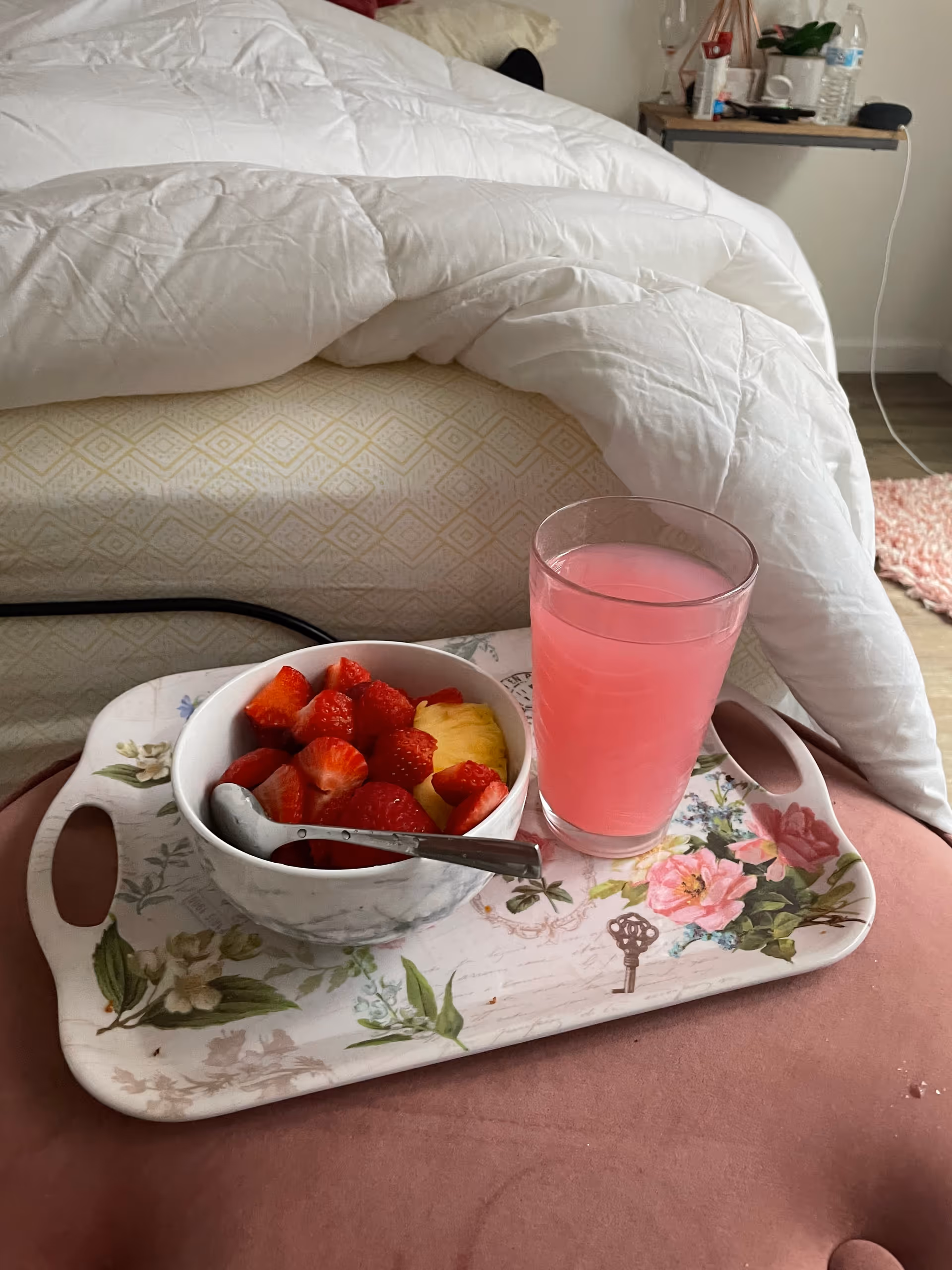 Bedside tray with a bowl of fruit and pink juice on a white quilted bed.