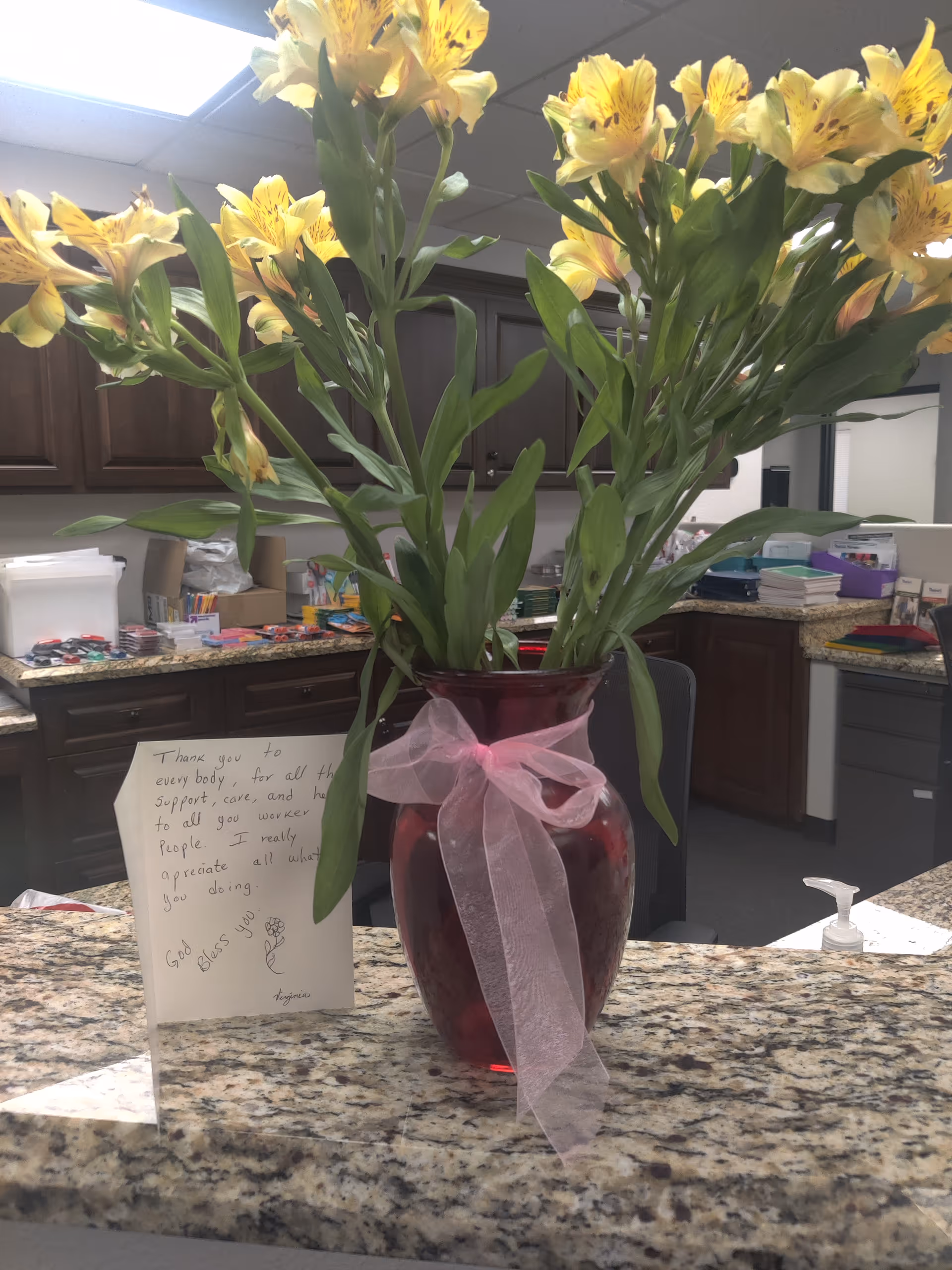 Yellow lilies in a magenta vase with ribbon sit on a granite counter in kitchen area.