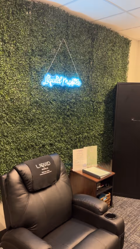 Indoor lounge area featuring a green leafy backdrop, neon Liquid Mobile sign, and a black recliner.