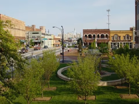 Outdoor city park and street with trees and brick storefronts.