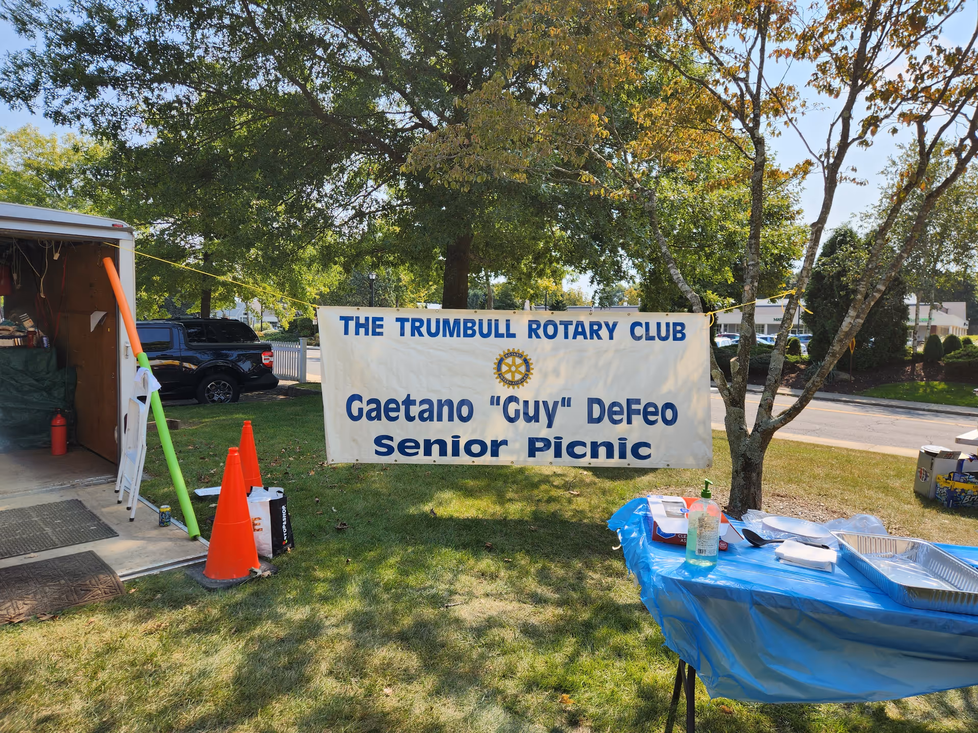 Outdoor senior picnic setup with banner, blue tables, cones, and a trailer on grass.