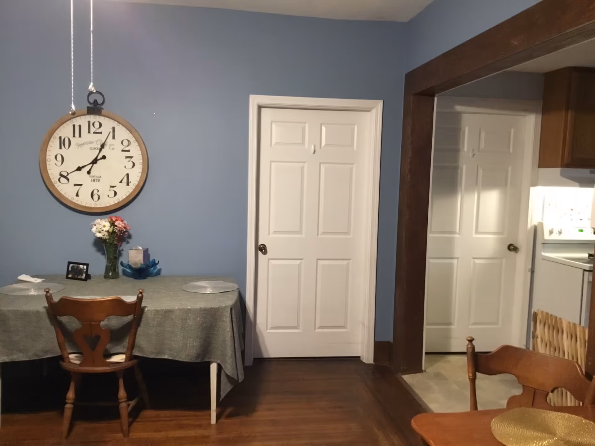 Blue-walled dining area with a large clock, table, chairs, and flowers.