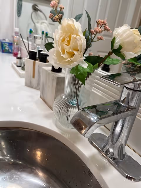 Bathroom sink area with chrome faucet, vase of white flowers, and grooming items on the counter.