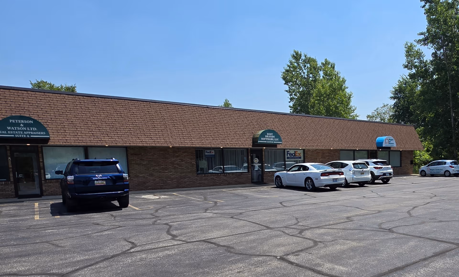 Single-story brick strip mall with storefronts and parked cars in parking lot.