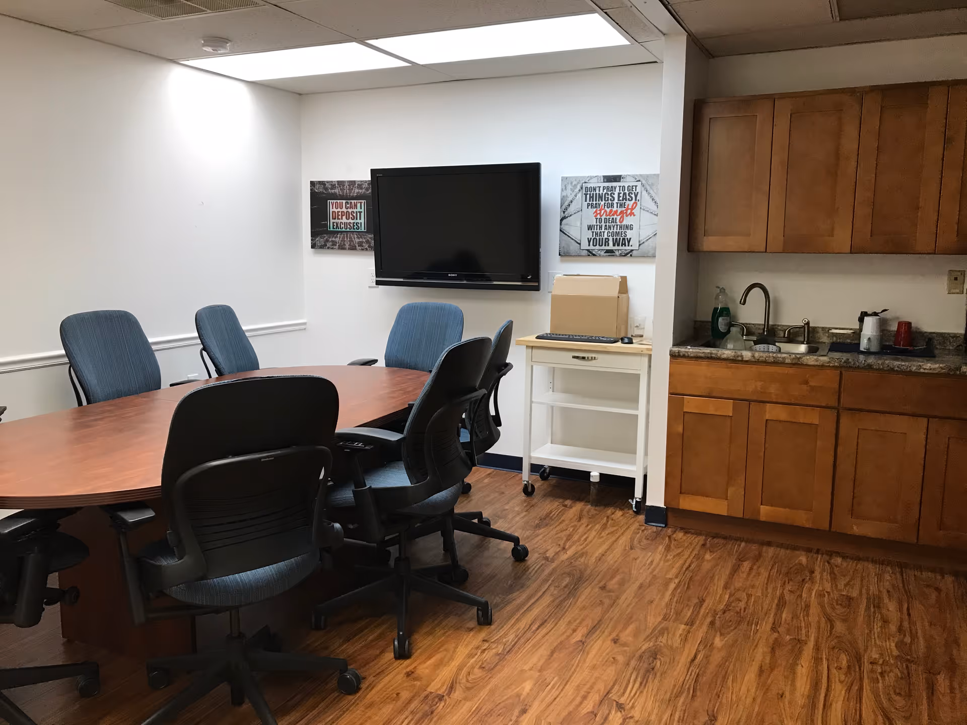 Conference room with oval table, blue chairs, wall-mounted TV, and kitchenette area.