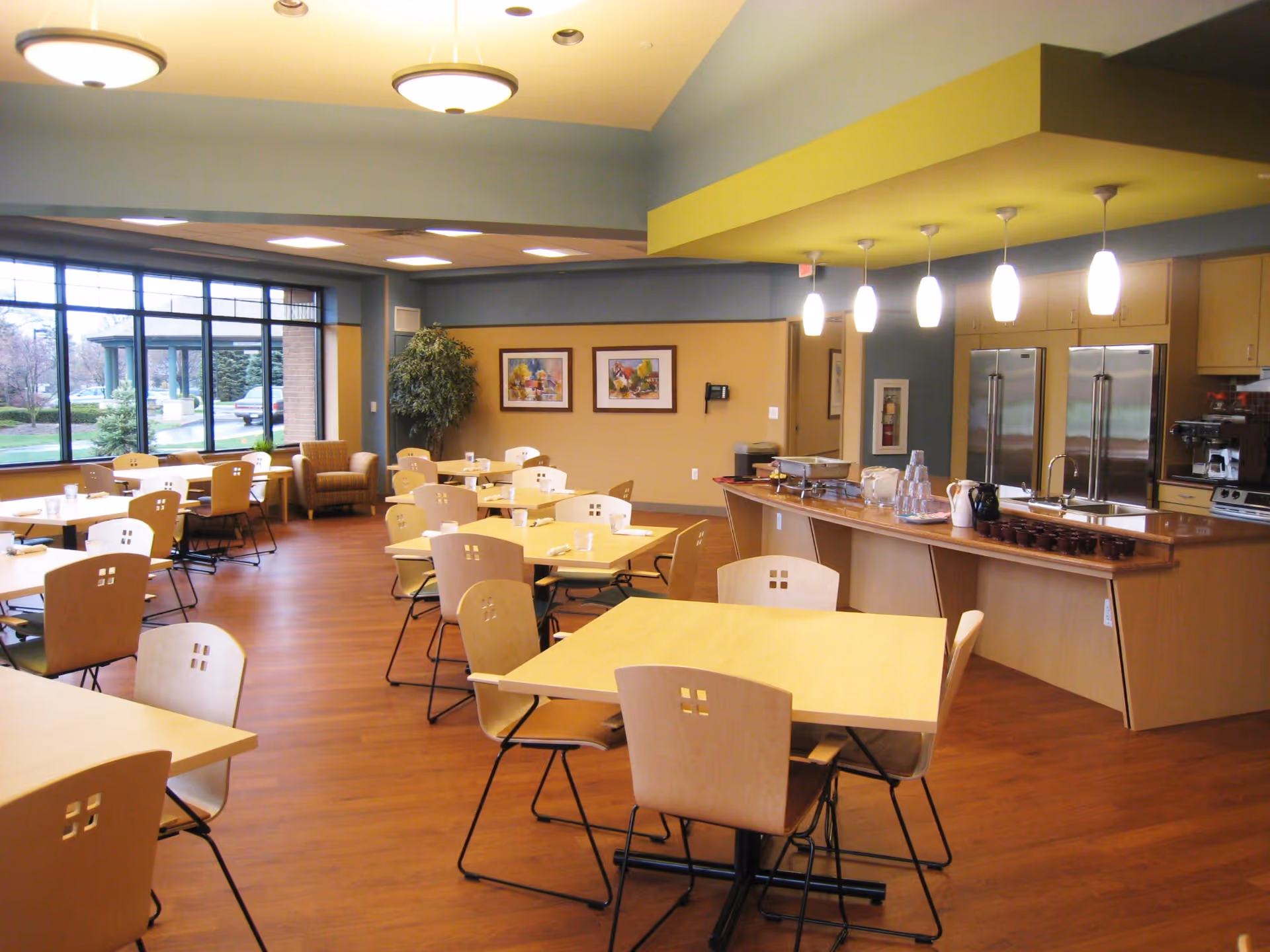 Bright senior living dining room with many tables, chairs, and a service counter.