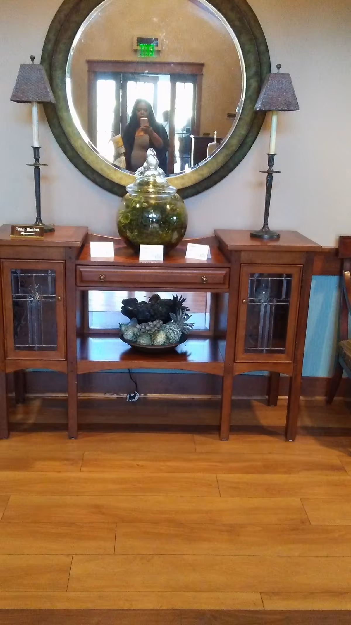 Wood console table with round mirror, lamps, ceramic jar, and a person reflected taking a photo.