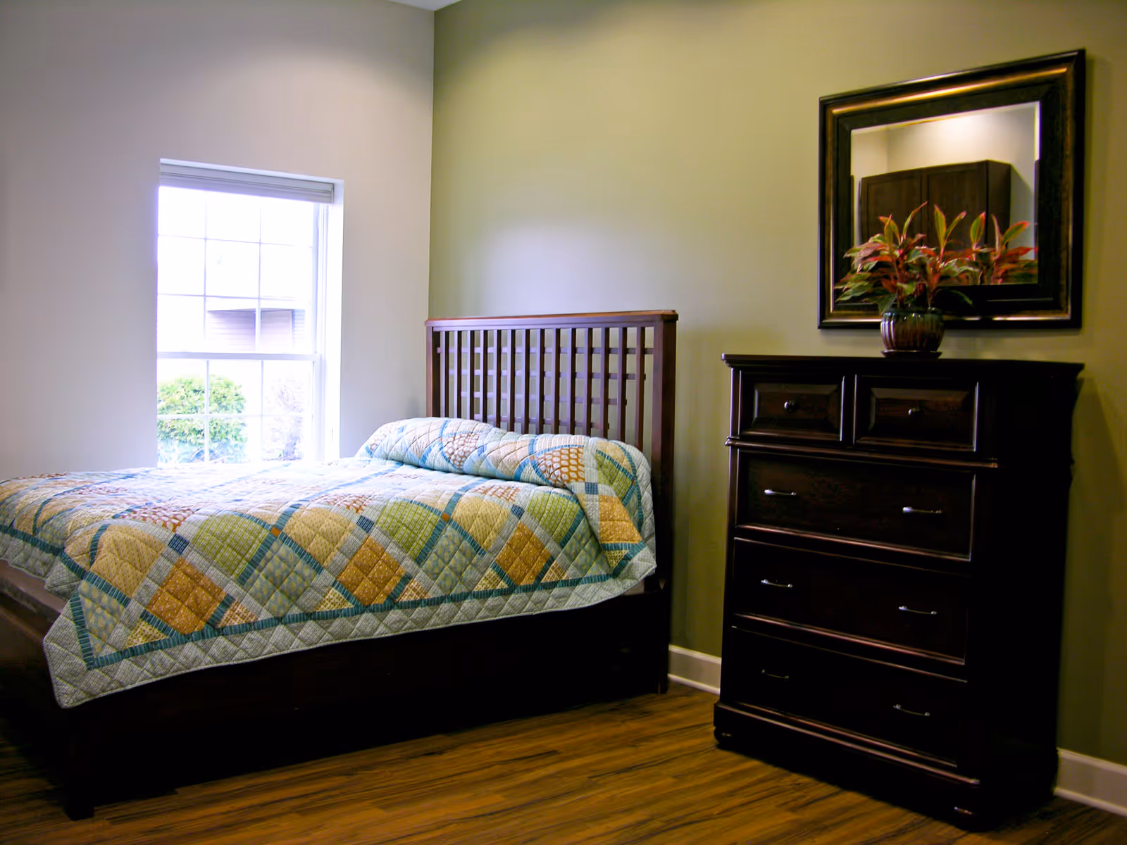 A tidy bedroom with a wooden bed, quilt, dresser, mirror, and green walls.