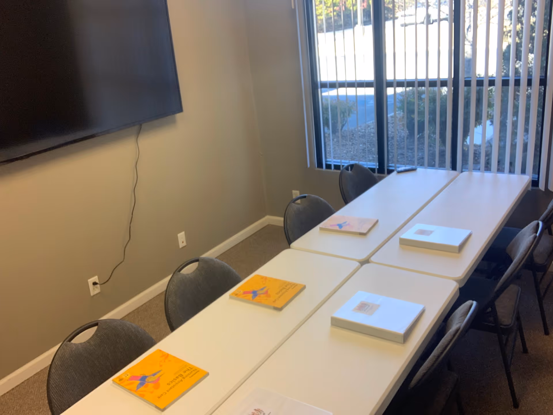 A conference room with white tables, chairs, a wall-mounted TV, and large windows.