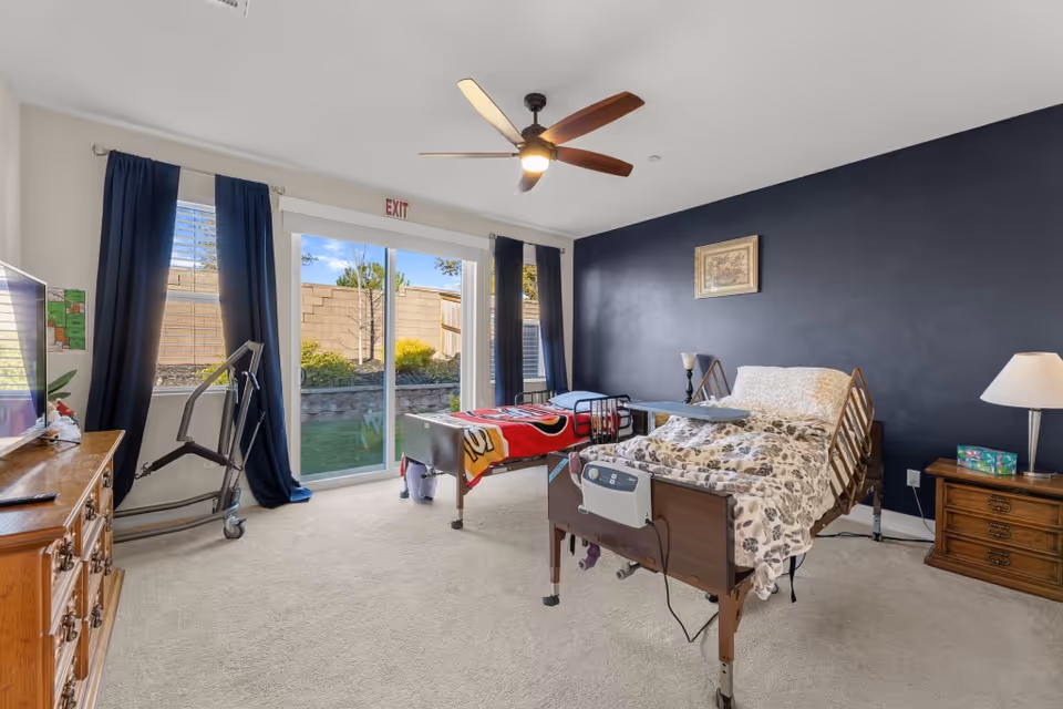 A furnished hospital bedroom with a bed, dresser, dark accent wall, and sliding glass door.