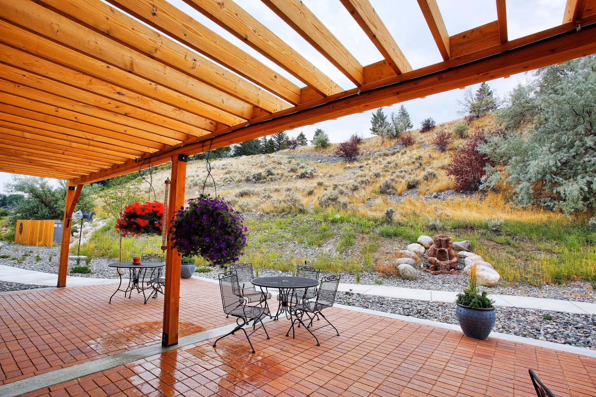 Outdoor covered patio with metal tables, hanging planters, and a hillside landscape.