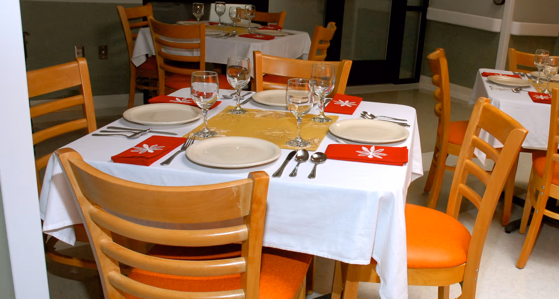 Empty dining room with white tablecloths, red napkins, and orange chairs.