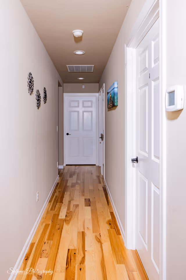 Bright, narrow hallway with hardwood floor, beige walls, and white doors in a senior living home.