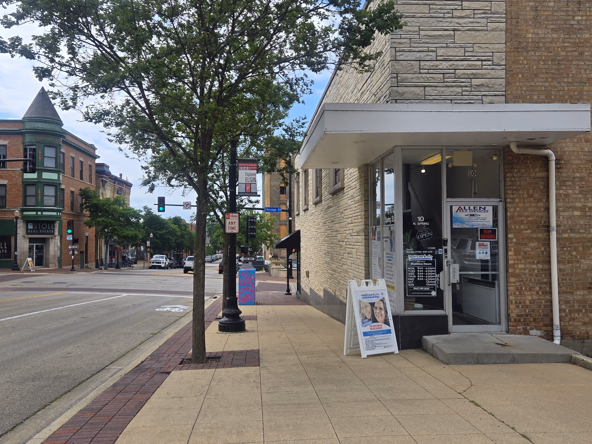 Urban sidewalk scene showing a brick building storefront, entrance, tree, and street traffic.