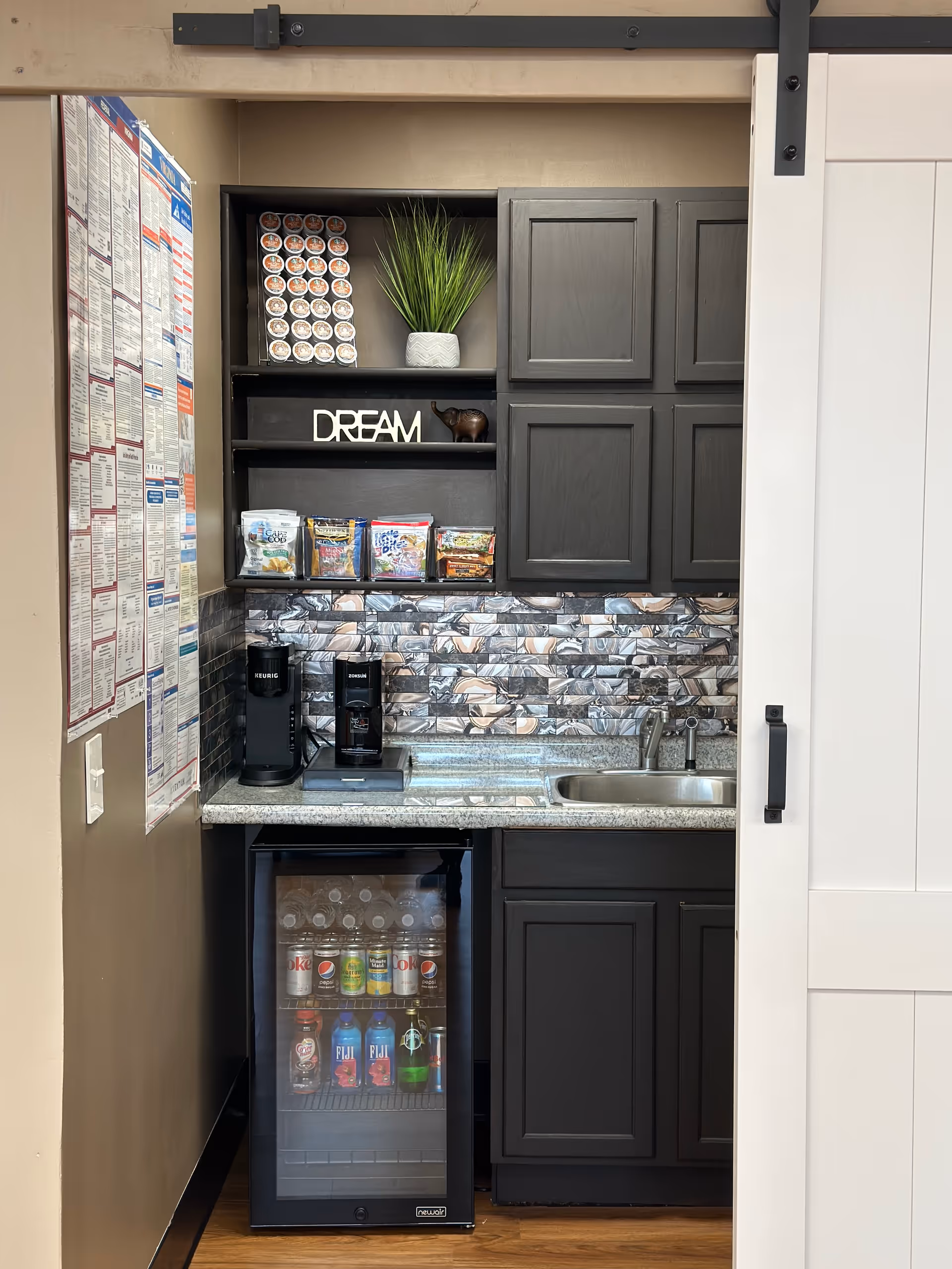 Small dark kitchen nook with coffee machines, snacks, drinks, and a sink.