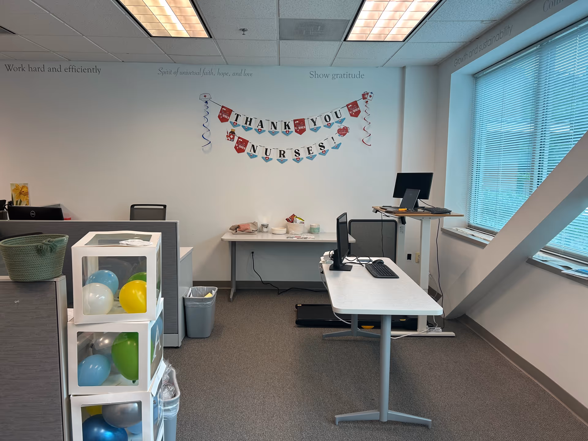 Interior office area with computer desk, celebratory banner, balloons, and large windows.
