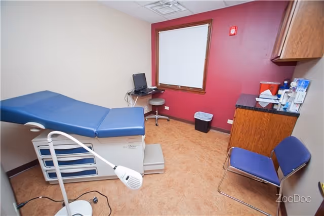 Medical examination room with blue padded exam table, computer, chairs, and cabinets.
