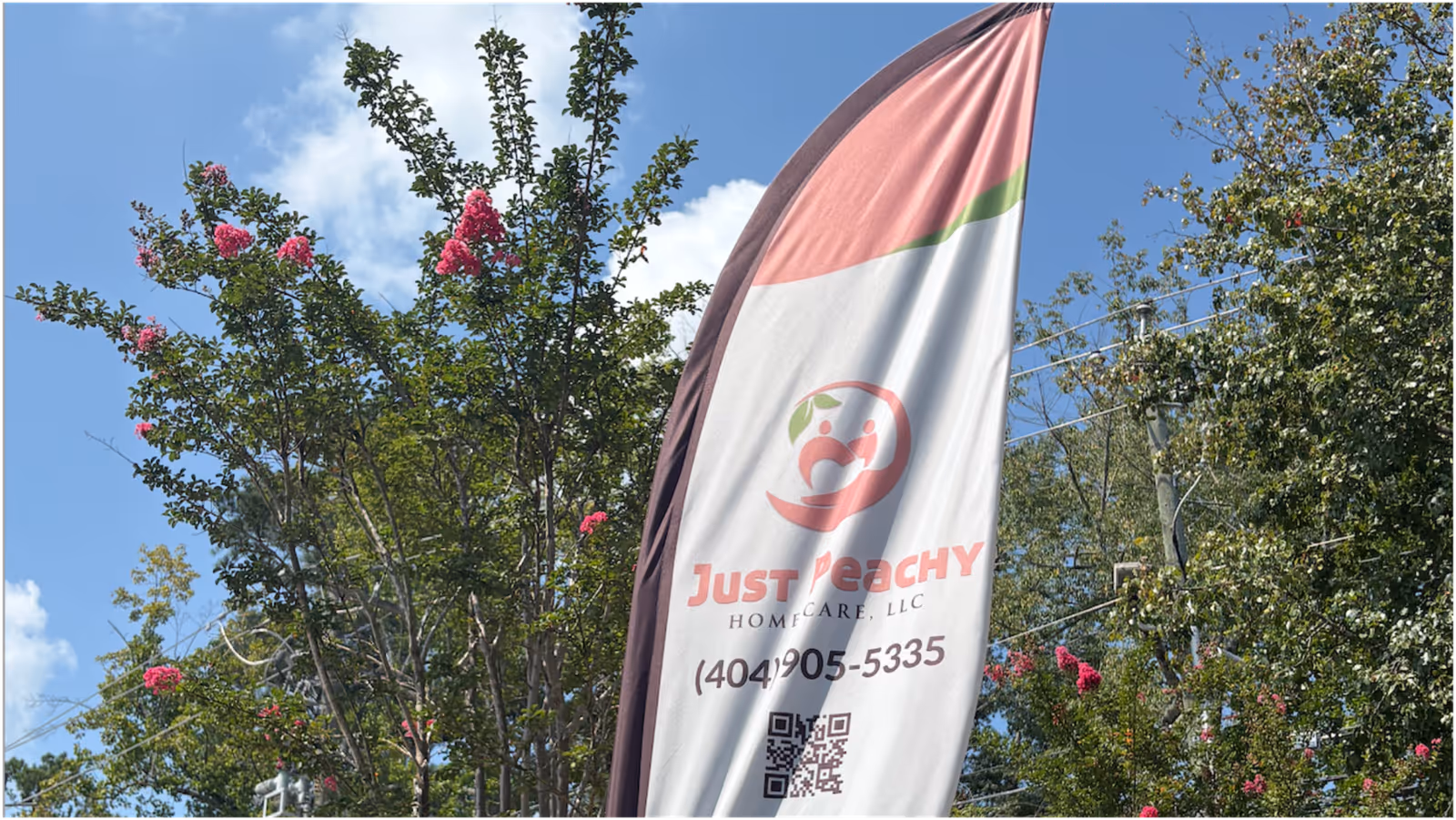 Outdoor scene with a tall banner advertising Just Peachy Home Care among trees.