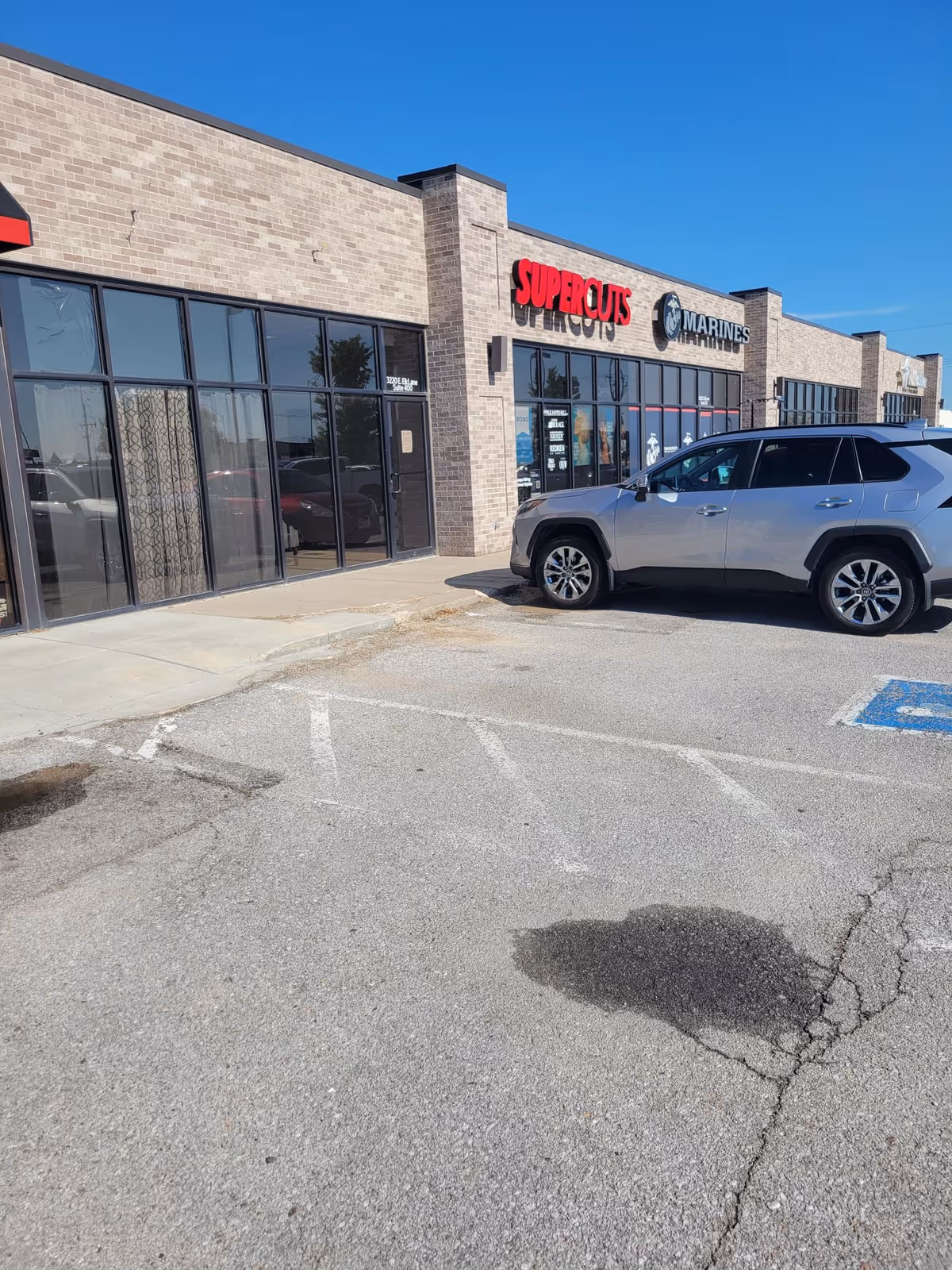 Facade of a strip mall with Supercuts sign and a parked silver SUV.