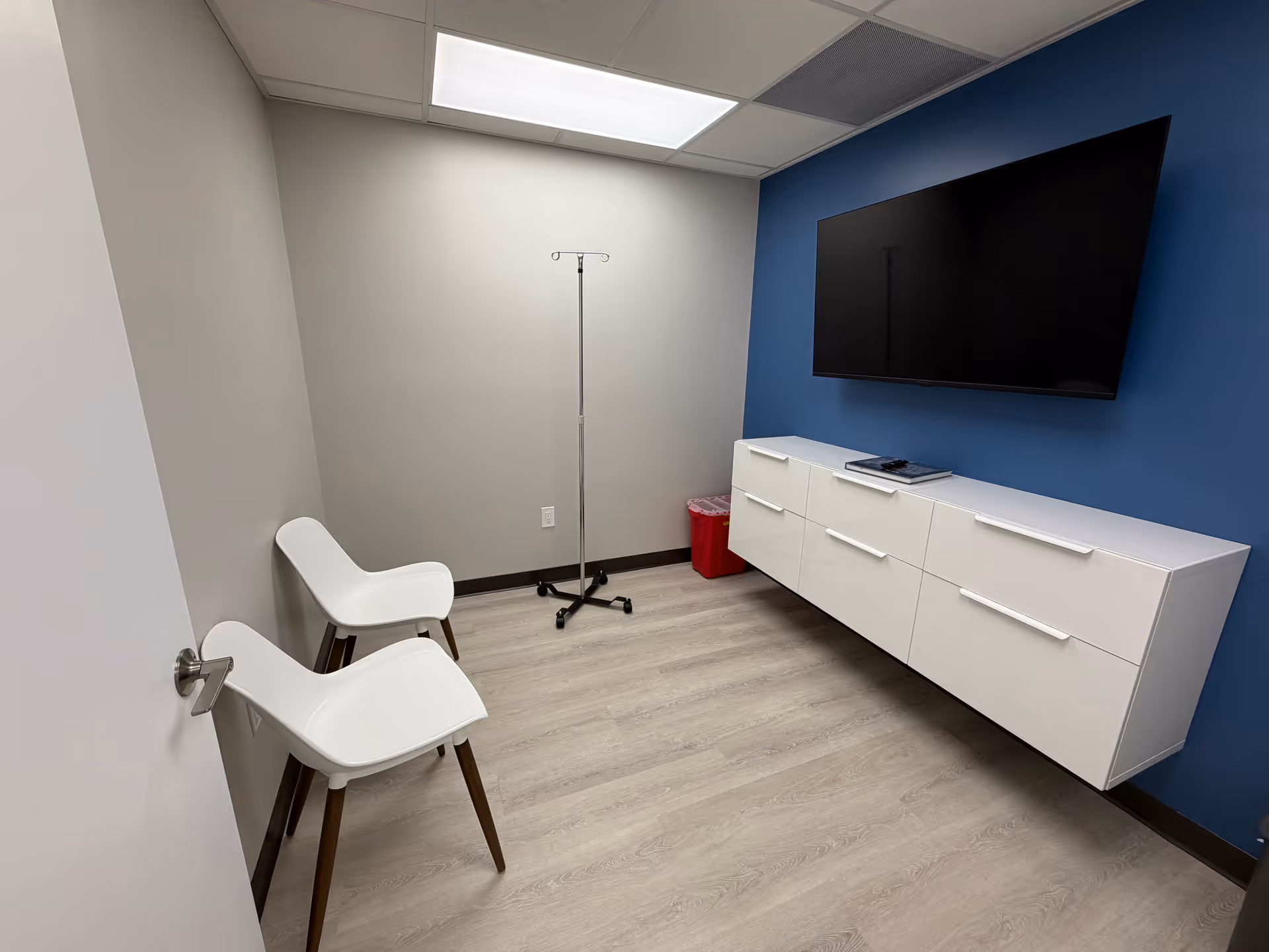 A clinical treatment room with two white chairs, an IV pole, and a wall-mounted TV.