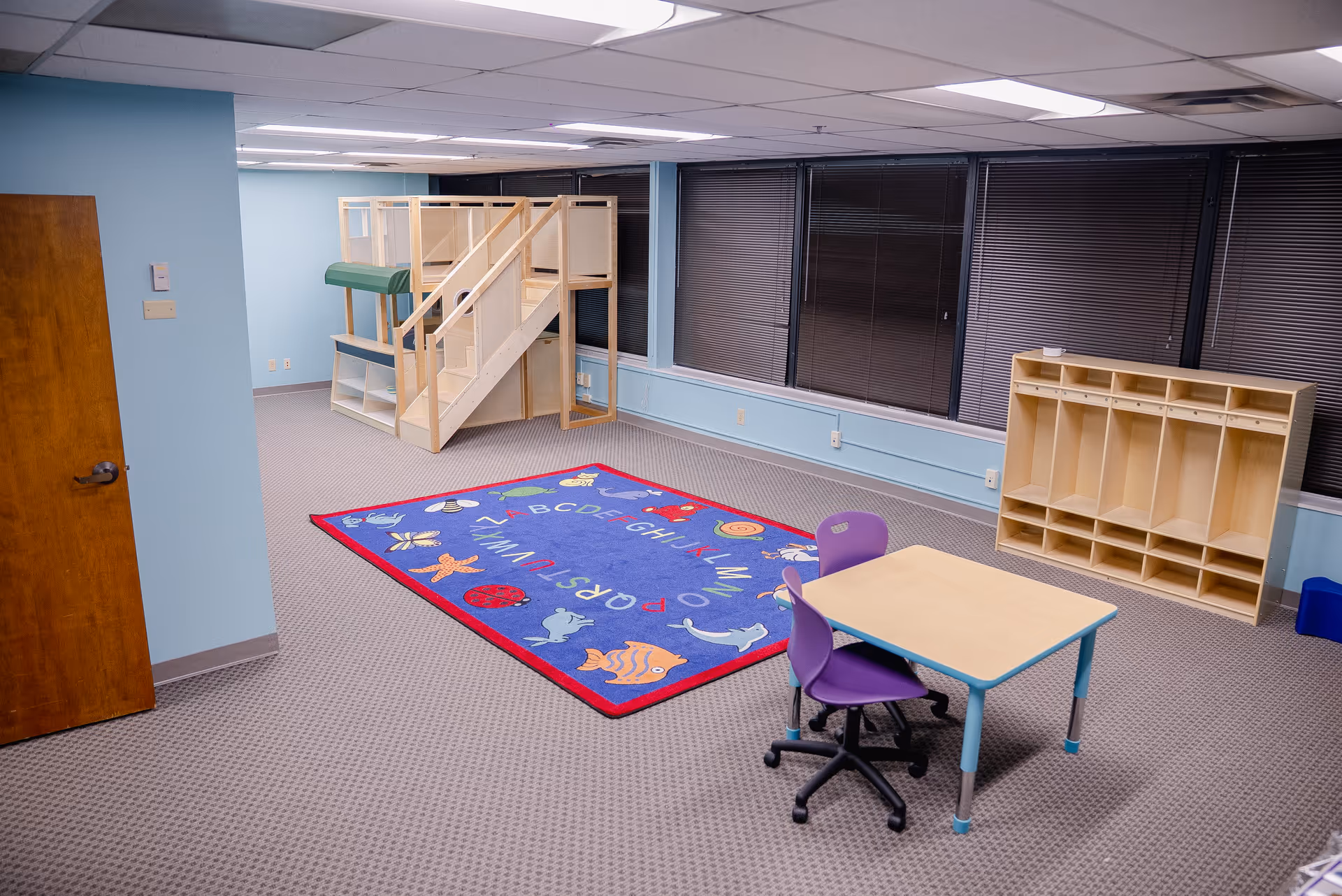 A bright indoor playroom with a wooden loft, alphabet rug, table, chairs, and cubbies.