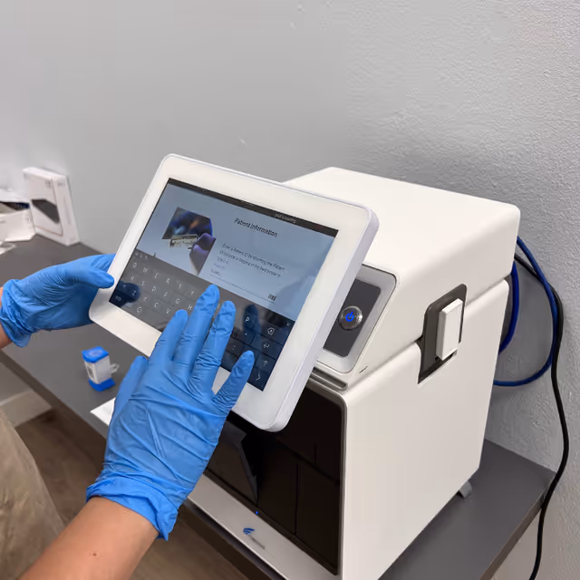 Gloved hands operate a tablet beside a white medical device on a clinical workstation.