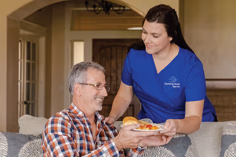 Caregiver in blue scrubs serves a plate to a smiling resident on a couch.