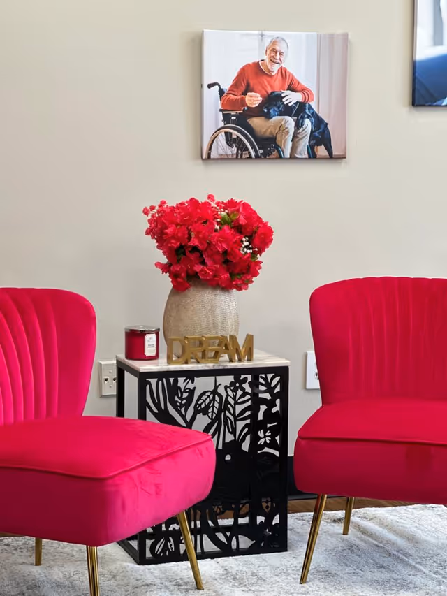 Bright pink chairs surround a black floral side table with a vase of red flowers.