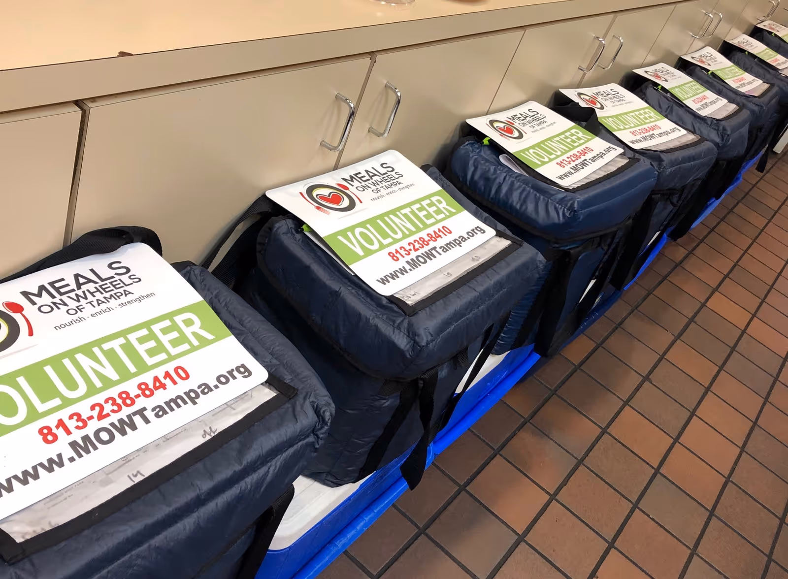 Row of volunteer bags labeled Meals On Wheels of Tampa along a counter.