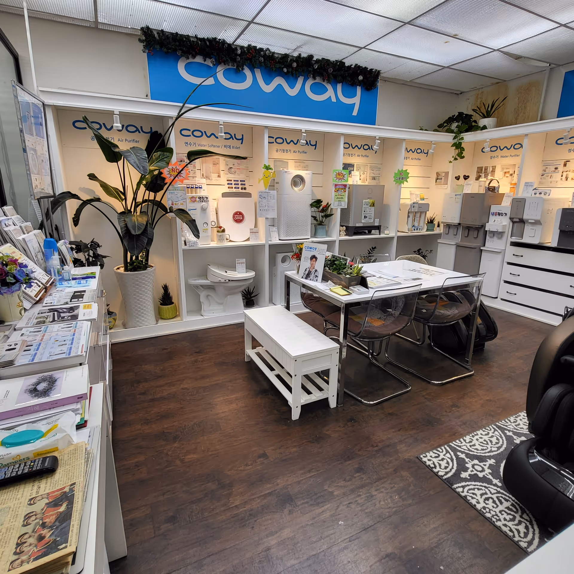 Showroom interior with Coway appliances, display shelves, table and chairs, potted plants, wooden floor.