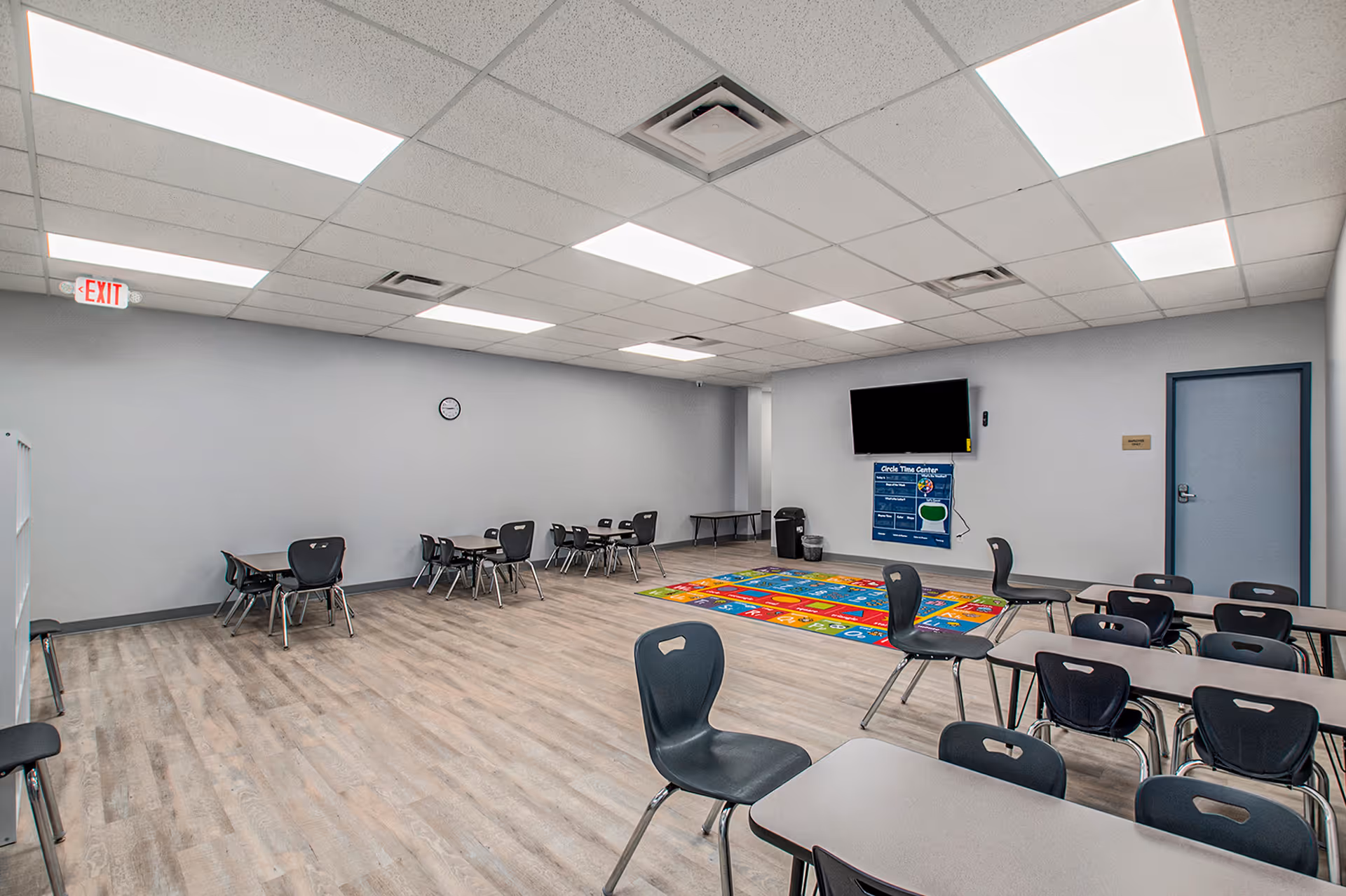 Bright multipurpose room with tables, chairs, a colorful rug, and wall-mounted TV.