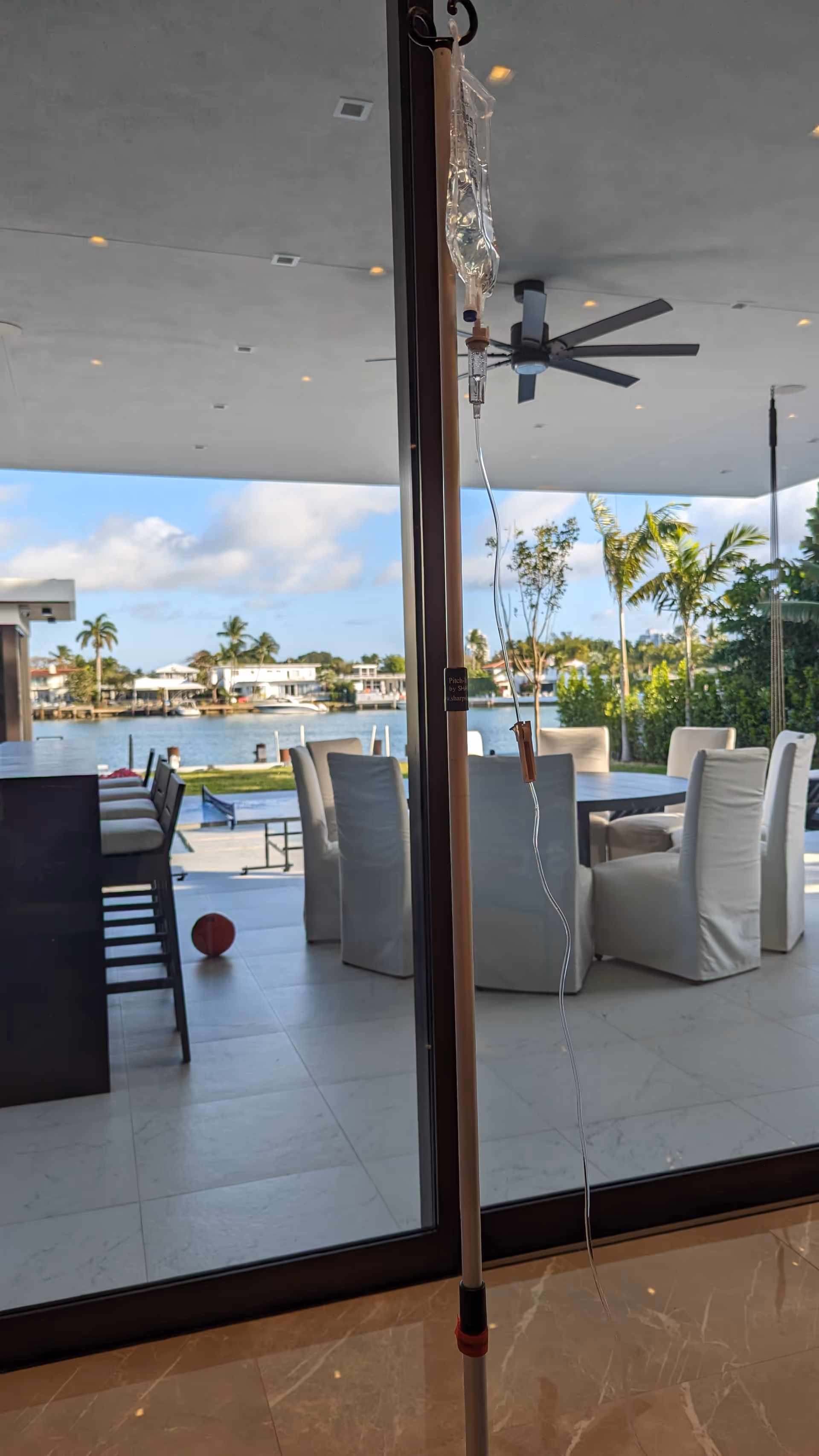 Outdoor seating area beside water is viewed through a doorway with an IV stand indoors.