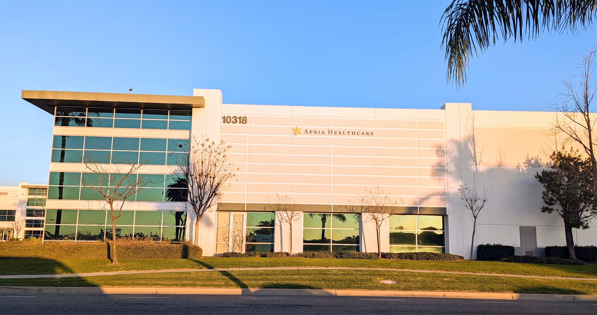 Modern white building labeled Apria Healthcare with large glass windows and palm trees outside.