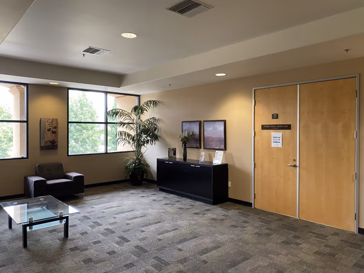 Spacious indoor lobby with a glass coffee table, black reception desk, large windows, and a plant.