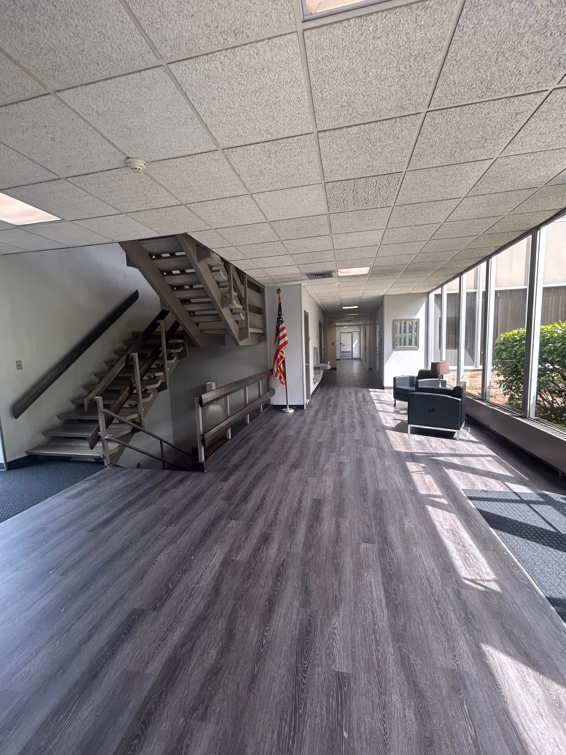 Long indoor hallway with staircase, American flag, and seating beside large windows.