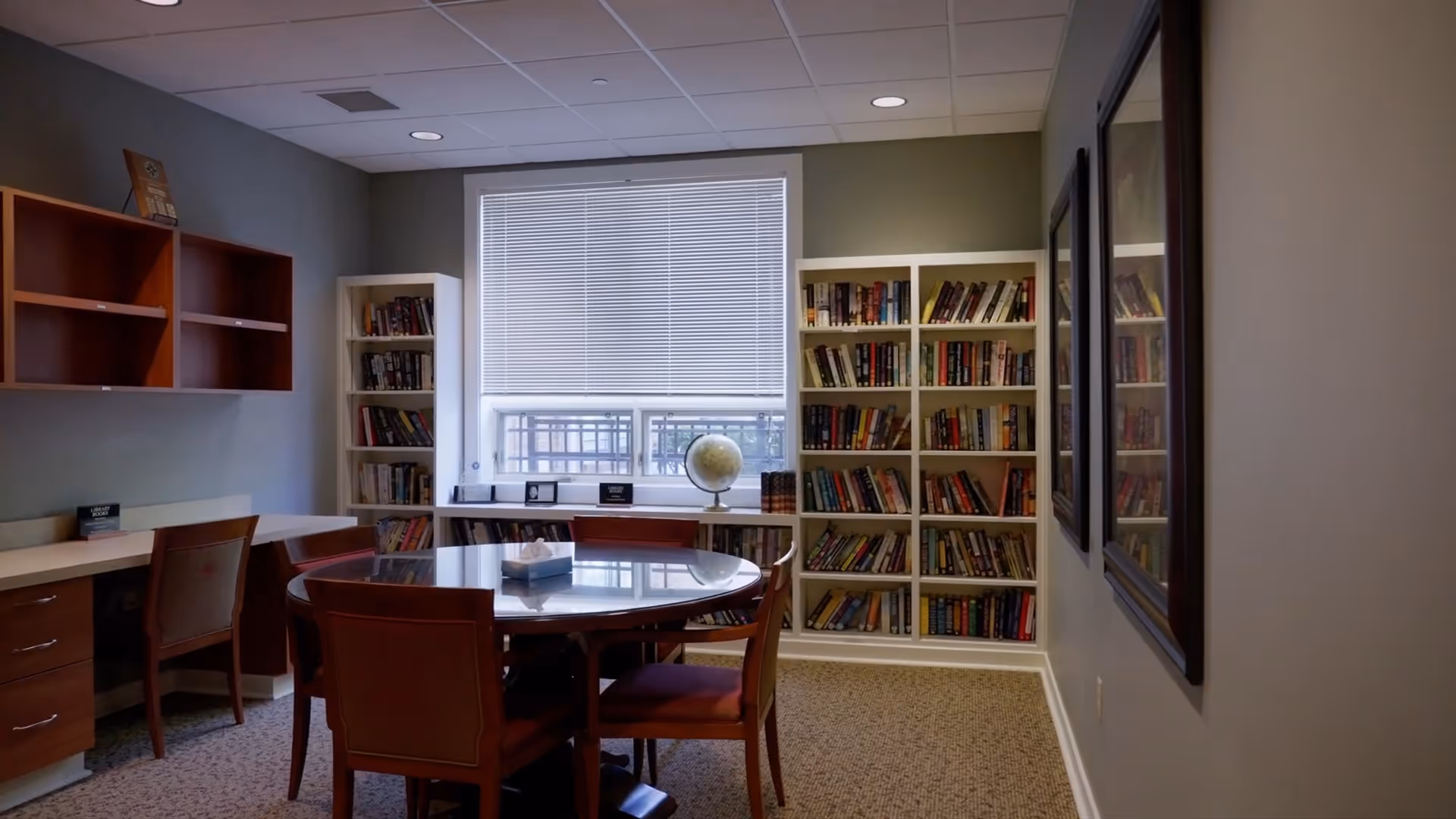 A quiet library-like room with bookshelves, a round glass table, and chairs by a window.