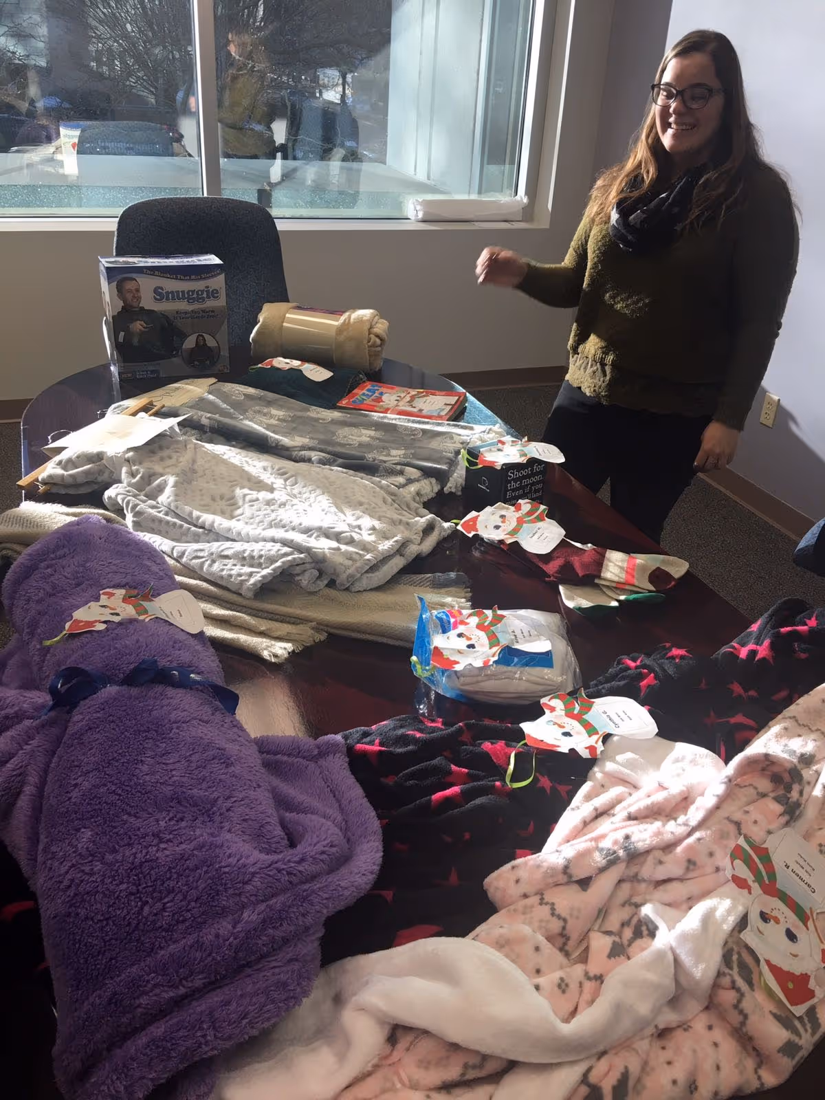 A staff member stands by a table stacked with blankets and clothing in a bright room.