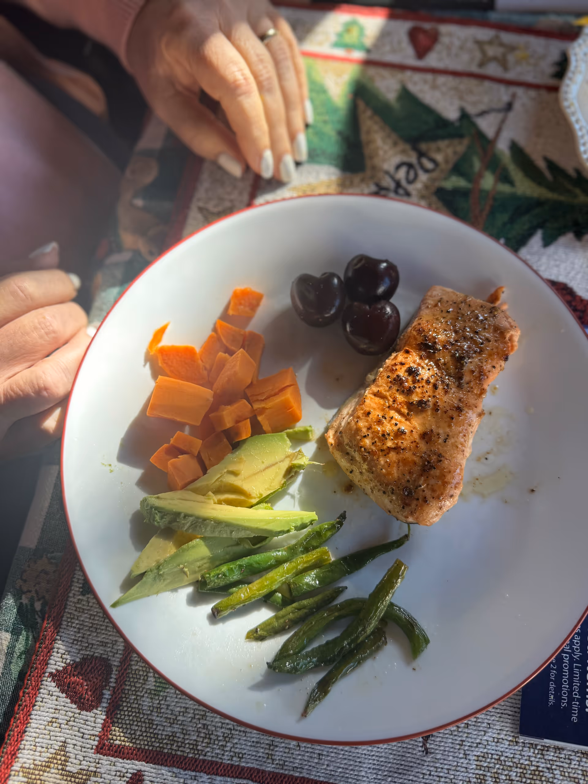 Plate with seasoned salmon, sweet potatoes, avocado, asparagus, olives; hands near the meal on a table.