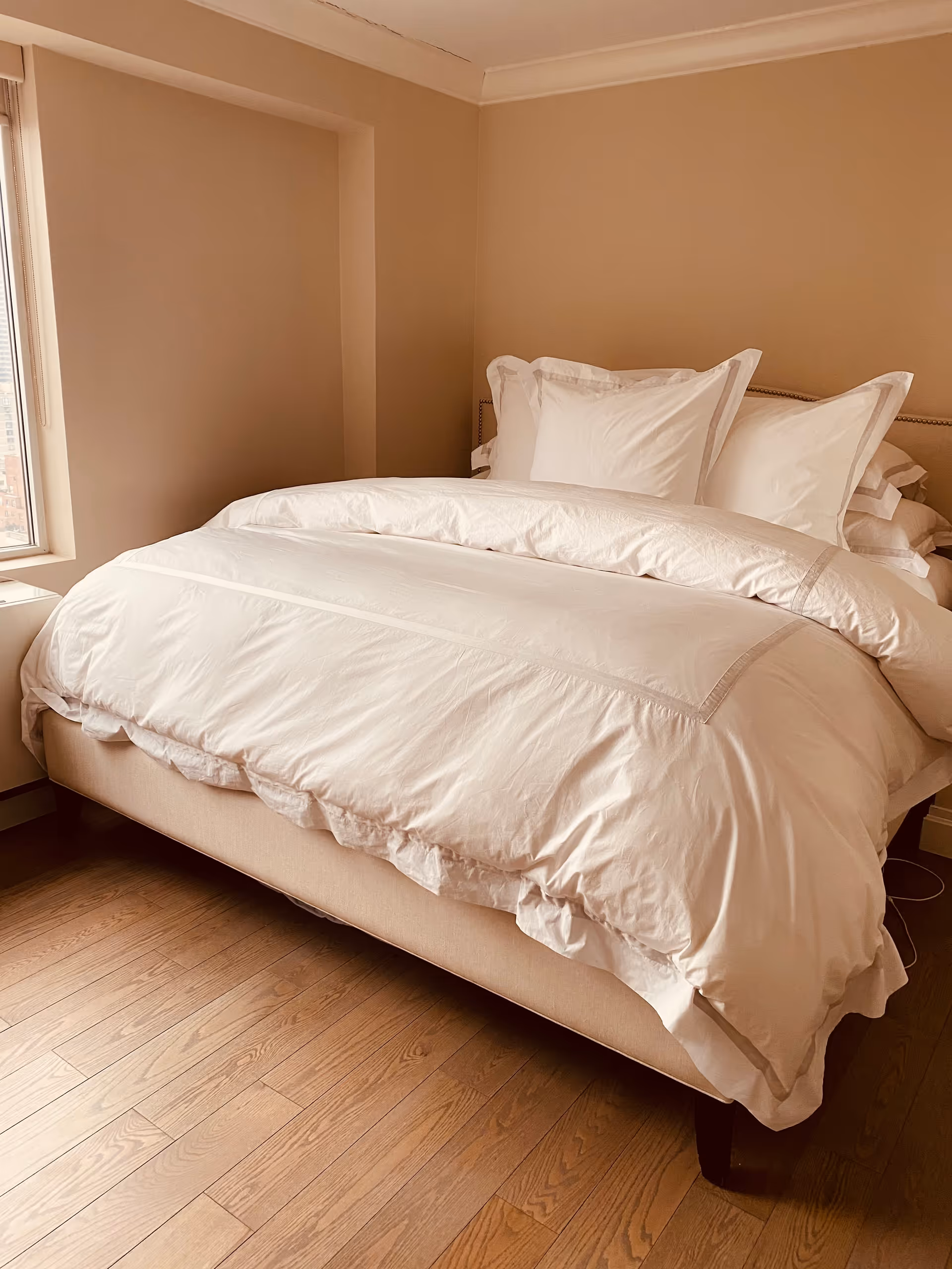 A neatly made white bed in a small beige bedroom with a window.