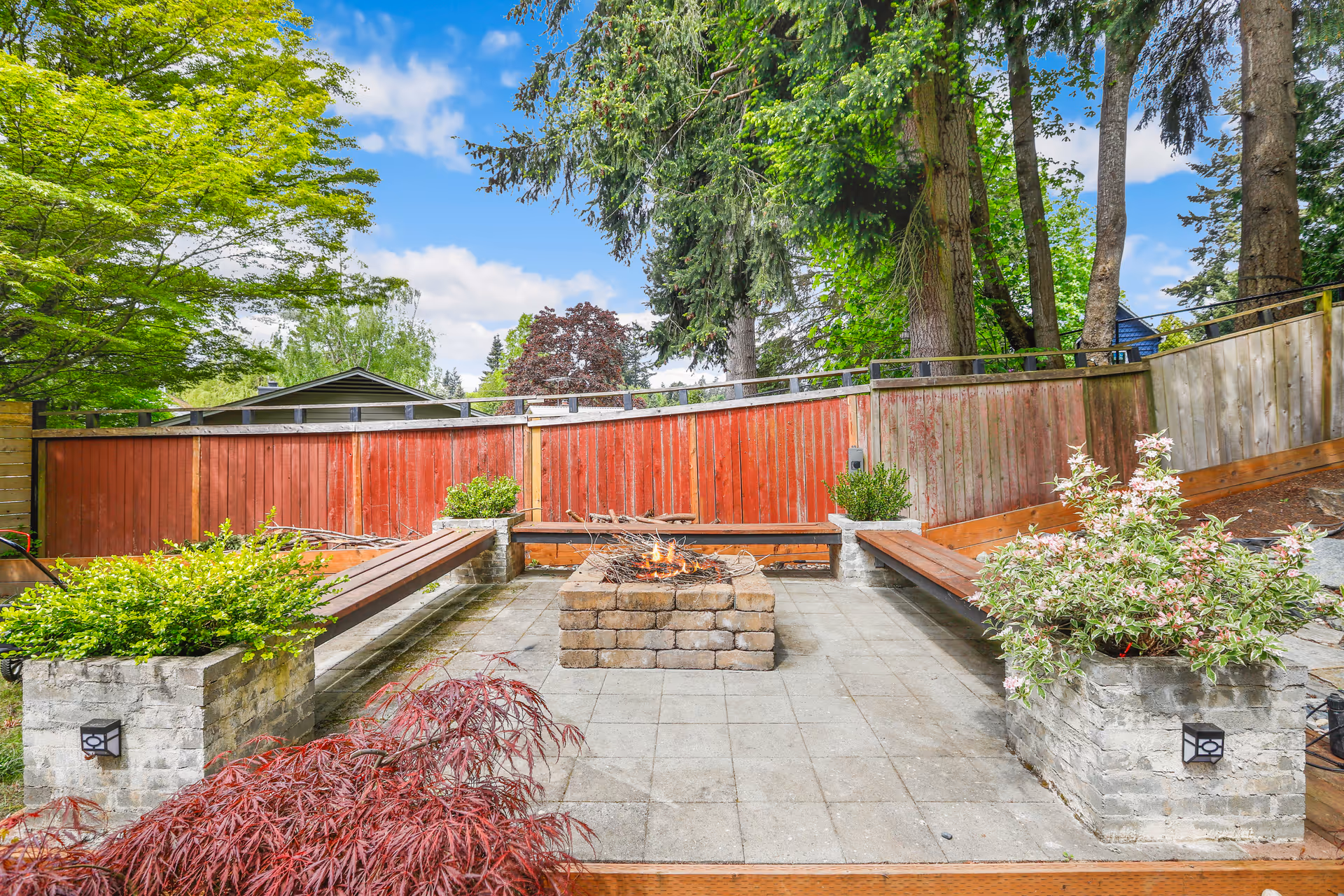 Outdoor courtyard with brick fire pit, benches, planters, fence, and trees.