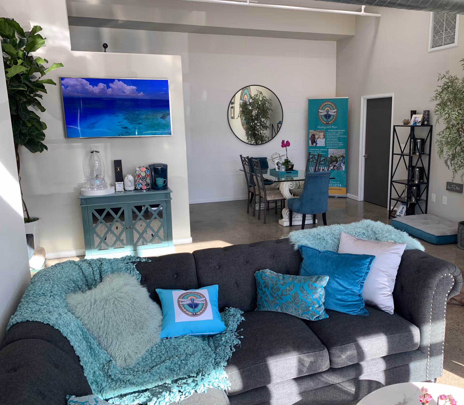Modern senior living lounge with gray sectional, teal pillows, and a dining area in the background.
