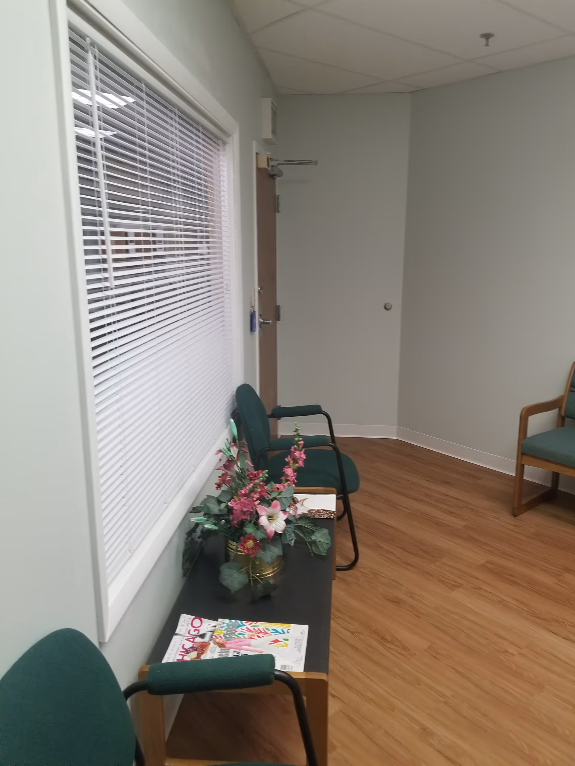 Interior waiting area with green chairs, window blinds, flowers, and magazines on a table.