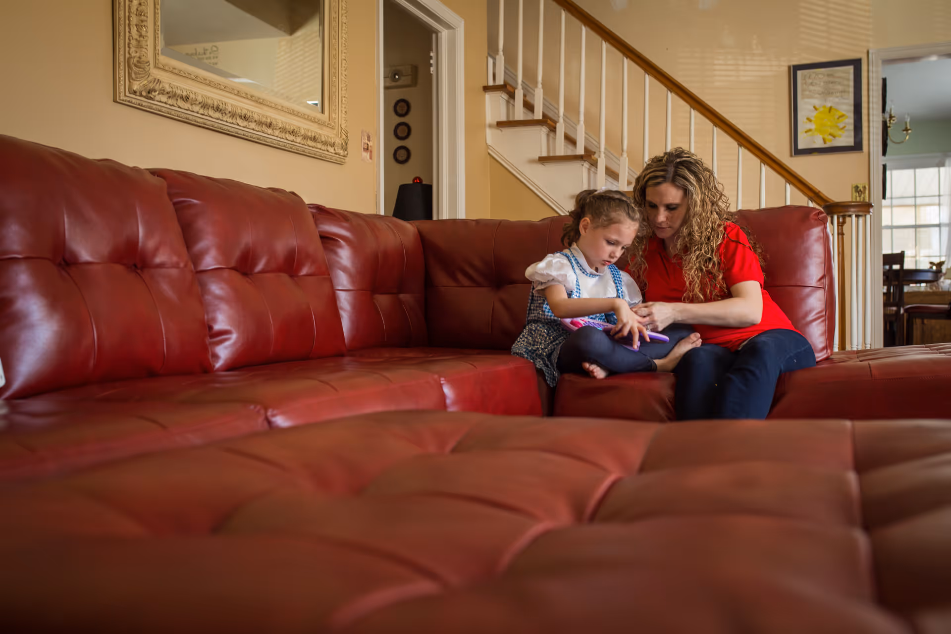 A woman and child sit on a red leather couch in a living room.